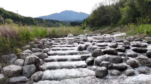 Clear water flows over smooth stones. Rocks form gentle cascades in the stream. Green grass and trees frame the scene. Mountains stand in the distant background. Nature's quiet energy fills the frame