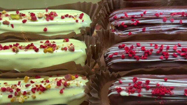 Close-up assorted eclairs on display with glossy chocolate and pale vanilla glazes, sprinkled with bright freeze dried raspberry