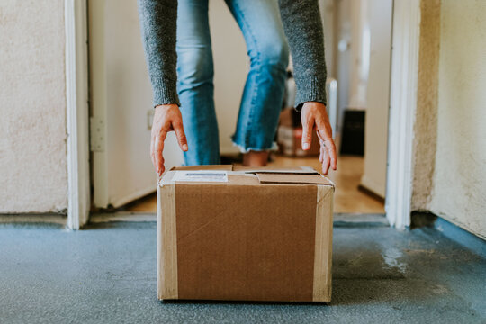 Person picking up a cardboard package from the doorstep, wearing jeans and a sweater.