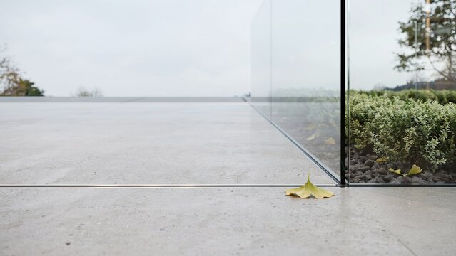 Glass partition divides a smooth concrete floor from a small patch of green hedges under a cloudy sky.