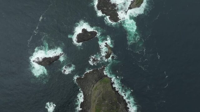 A push in top down aerial drone shot captures the dramatic, remote coastline and sea stacks on the Faroe Islands. Waves crash against the cliffs near Ei&eth;iskollur mountain, showcasing raw nature.