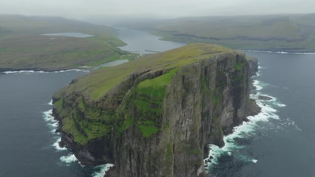 Aerial drone shot panning left, capturing the majestic Ei&eth;iskollur mountain and the village of Ei&eth;i on the Faroe Islands. Overcast skies highlight the rugged cliffs, green landscape, and vast ocean.