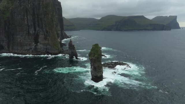 A breathtaking aerial pan left tracking shot of the rugged coastline and towering Ei&eth;iskollur mountain on the Faroe Islands, featuring dramatic sea stacks amidst a misty, cloudy day.