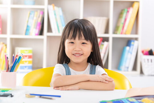 A polite and happy little Asian girl sitting up straight at her desk with crossed arms and smiling brightly at the camera.