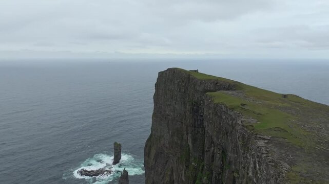 Push in aerial drone shot showcasing the rugged coastline and majestic cliffs of Ei&eth;iskollur mountain on the Faroe Islands. A remote, wild landscape under cloudy skies.