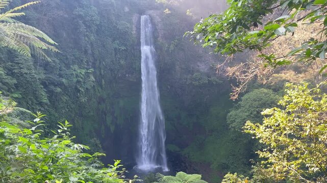 shot of a tall tropical waterfall cascading down a mossy cliff surrounded by dense green rainforest vegetation in West Java, Indonesia. Water flows continuously with natural motion blur. Peaceful natu