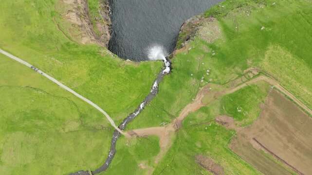 Aerial view of the spectacular M&uacute;lafossur waterfall falling into the ocean on the Faroe Islands. Featuring lush green landscape, rugged cliffs, and a hiking trail.
