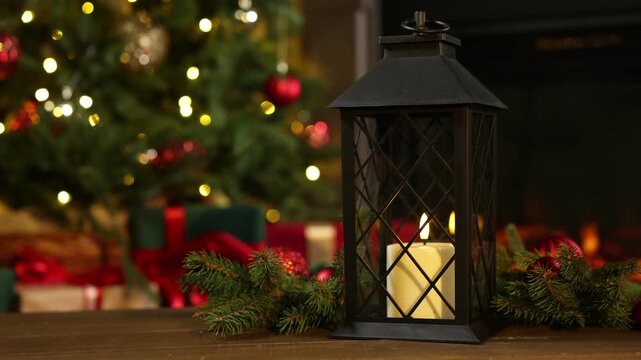 Christmas lantern with burning candle, fir tree branches and baubles on wooden table indoors against blurred lights and fireplace, closeup