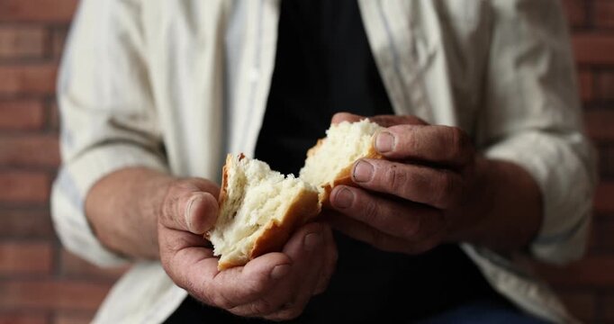 Poverty. Homeless senior man breaking piece of bread indoors, closeup. Camera moving forward