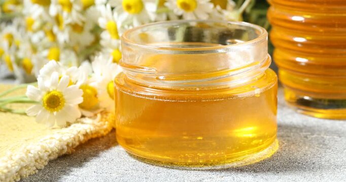Woman putting chamomile flowers and dipping spoon into jar with sweet honey at grey table, closeup