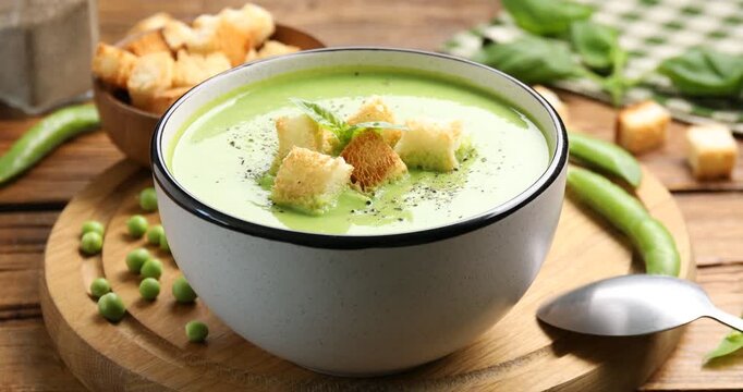 Woman adding basil to delicious pea cream soup with croutons at wooden table, closeup