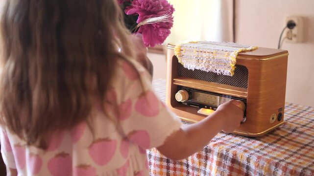 Young girl adjusting the knob of a vintage wooden radio, feeling a sense of nostalgia while surrounded by blooming peonies on a checkered tablecloth