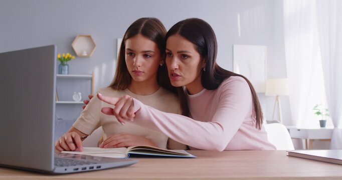 Supportive mother helping small girl with homework embraces her with love andd explains something carefully, indicates at monitor of laptop computer. Family doing school task together