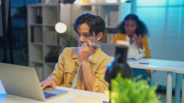 asian male analyst using AI agent on laptop for business planning at night office with colleagues working in background, artificial intelligence assistant and corporate strategy concept