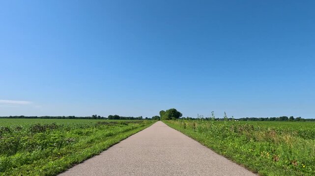 Slow-motion view from a moving bicycle receding down a paved recreational trail passing between lush green, healthy soybean fields under a clear blue sky.