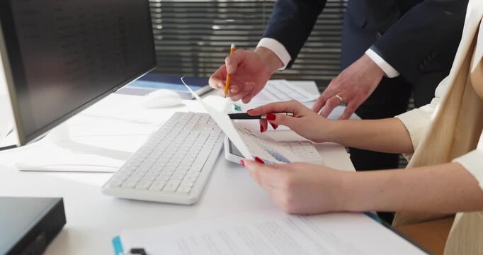 Colleagues review finance paperwork with charts in office. Hands point to accounting near keyboard and computer as numbers are checked and notes added. Clear office business teamwork concept.