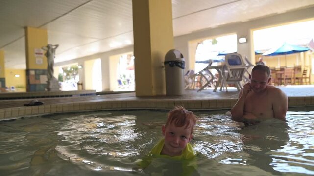 Young boy in bright swimwear splashes in the pool while an adult male observes, both engaged in playful interaction on a sunny day at a resort with lounge chairs and umbrellas in the background