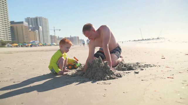 Caucasian father and son engage in playful activity on a sandy beach, constructing a sandcastle with bright toys, while tall buildings and a sunny sky create a vibrant coastal atmosphere