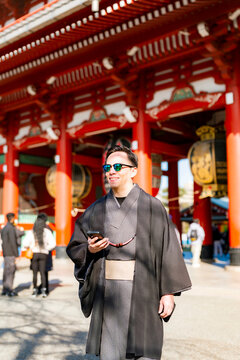 Young smiling Latin man walking confidently while holding his phone at Sensoji temple in Tokyo Japan, wearing a traditional dark kimono or yukata with elegant style, surrounded by pagoda and daylight