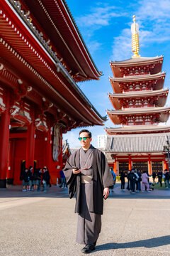 Young smiling Latin man walking confidently while holding his phone at Sensoji temple in Tokyo Japan, wearing a traditional dark kimono or yukata with elegant style, surrounded by pagoda and daylight