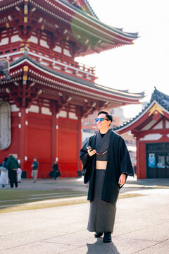 Young smiling Latin man walking relaxed through a temple corridor at Sensoji in Tokyo Japan, wearing a dark traditional kimono or yukata with elegant style, surrounded by vivid red wooden architecture