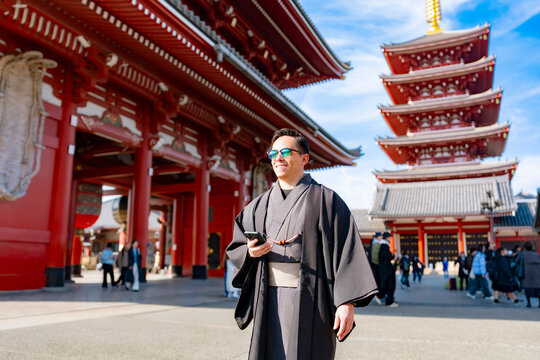 Young smiling Latin man walking confidently while holding his phone at Sensoji temple in Tokyo Japan, wearing a traditional dark kimono or yukata with elegant style, surrounded by pagoda and daylight