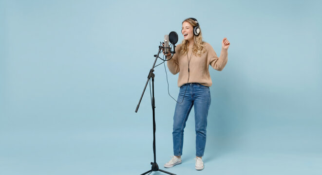 A woman stands next to a microphone in a recording studio. She wears headphones and sings with a joyful expression. The background is light blue and she is dressed in casual clothes