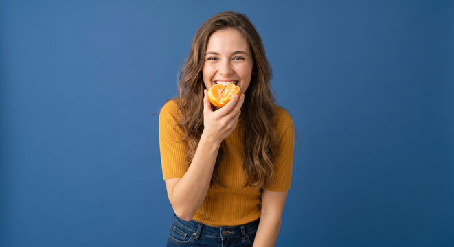 A woman stands in front of a blue wall holding a slice of orange. She smiles widely while enjoying the fruit. Sunlight fills the studio, creating a bright atmosphere