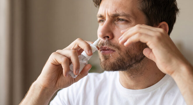 A man is using a nasal spray while sitting in a bright indoor area. He is focused on his action and appears to be in a personal space. Sunlight shines in through a window