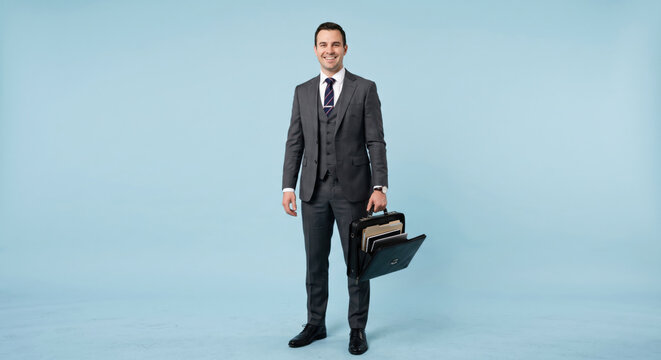 A man in a suit stands smiling while holding a briefcase in a studio with a light blue background. He appears ready for a business meeting or event