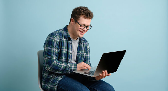 A young man sits on a chair with a laptop in his lap. He is focused and smiling while working. The background is light blue. It looks like a home office setting