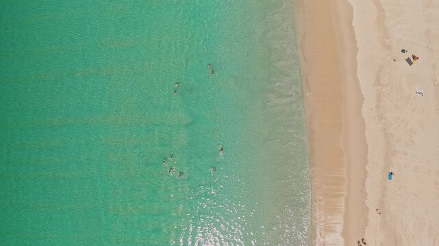 Clear turquoise ocean water sparkles as people swim near the sandy shore, enjoying a relaxing tropical vacation on Phuket's popular beach in Thailand from an aerial perspective, top down view