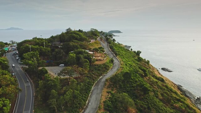 Promthep Cape, popular viewpoint on Phuket island, reveals a curving road along the lush tropical coastline with distant islands, boats traversing calm turquoise Andaman Sea. Aerial view drone flight