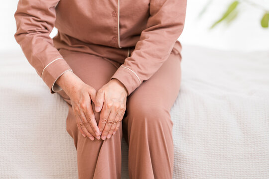 A senior woman in pajamas sits on her bed after waking up, complaining of knee pain (knee, knee pain).