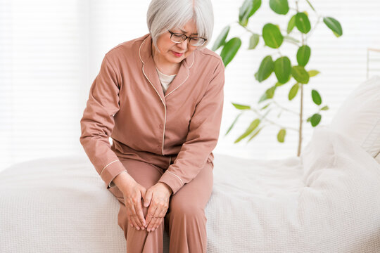 A senior woman in pajamas sits on her bed after waking up, complaining of knee pain (knee, knee pain).