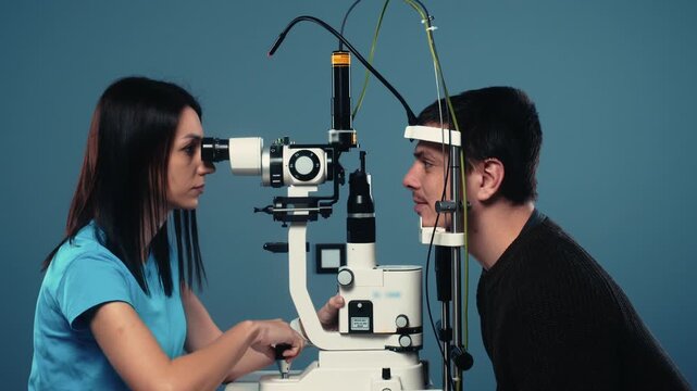 Female optometrist examines male patient's eyes using a slit lamp in a clinical setting with a blue background and medical equipment visible