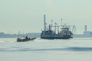 Fototapeta premium Ferry and small fishing boat crossing hazy harbour water near an industrial skyline at sunrise.