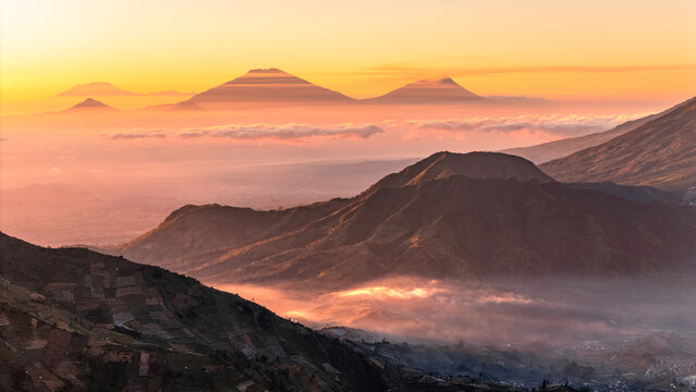 Aerial view of volcanic mountain peaks rising above a sea of mist during golden hour with terraced agricultural fields on the slopes in Dieng, Jawa Tengah, Indonesia.