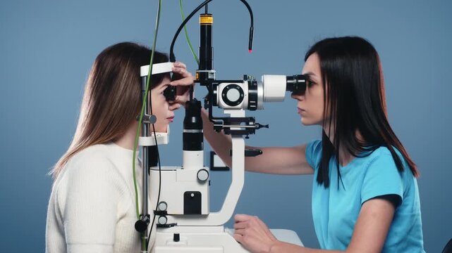 Female optometrist examines patients eyes using a slit lamp, showcasing detailed evaluation of vision in a clinical setting with blue background