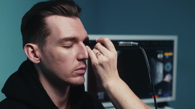 Male patient undergoing ultrasound examination of the eye with a handheld device, medical equipment visible in the background, clinical setting with blue walls
