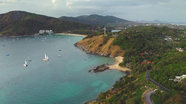 Nai Harn Beach in Phuket, turquoise water bay, sailboats anchored, green hills, and a wind turbine offering a scenic tropical landscape of Thailand coastline on a cloudy day. Aerial view drone flight