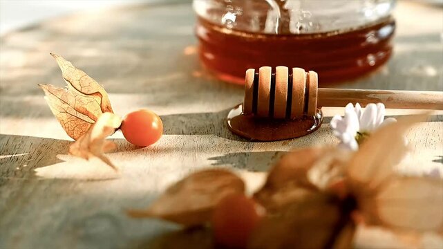Close-up flat lay of a wooden honey dipper with physalis husks and a daisy on a sunlit wooden board near a honey jar.