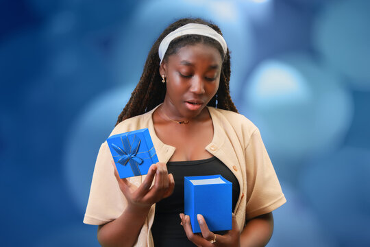 curious young Black woman opening a blue gift box against a blurred background