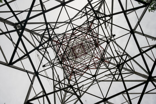 Abstract symmetrical view looking up through the steel lattice framework of a high-voltage power transmission tower against a pale sky. Repeating geometric beams illustrate the power grid energy