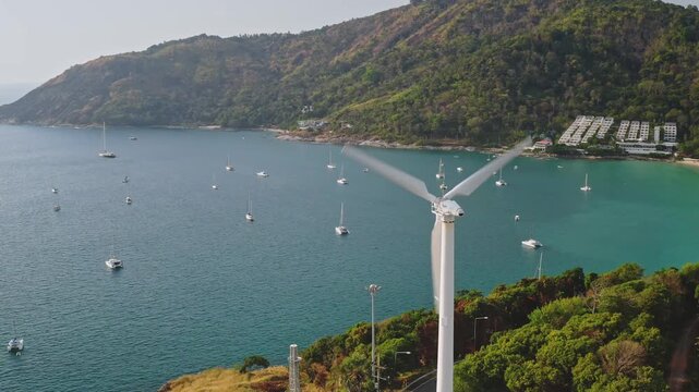 Aerial view panorama of wind turbine blades spinning, generating renewable energy on an island hill overlooking a tropical bay filled with sailboats, lush green coastline and beach in Phuket, Thailand