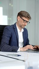 Fototapeta premium Professional architect or engineer actively working on a laptop. Analyzing architectural blueprints and building plans. Developing new projects in a modern office environment