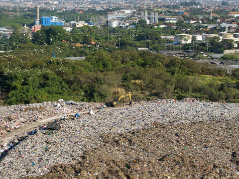 Aerial view of TPA Suwung landfill with an excavator working on waste piles, surrounded by lush vegetation and urban infrastructure in Kecamatan Denpasar Selatan, Bali, Indonesia.