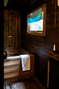 Rustic dark wooden soaking tub with a folded white towel and vintage brass shower in a forest cabin bathroom. A small window with a teal blind lets in soft daylight creating an intimate spa-like
