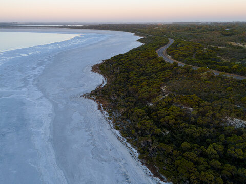 Aerial view of Pink Lake with salt crust shoreline, adjacent to native bushland and a winding coastal road under a soft twilight sky in Esperance, Western Australia, Australia.