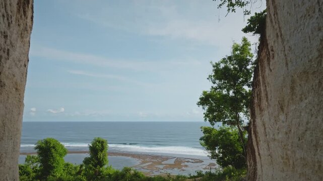 Drone flight between eroded limestone cliffs revealing Green Bowl Beach, its sandy shore, and the vast Indian Ocean with breaking waves under a clear blue sky on the tropical island of Bali, Indonesia